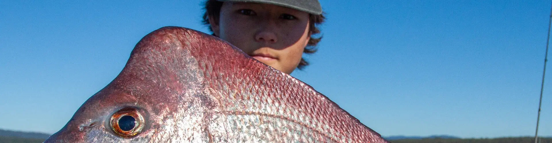 Young angler holding a pink snapper close to the camera against a blue sky