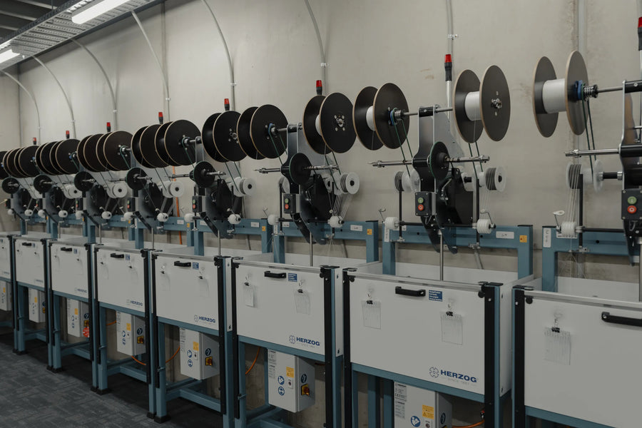 Row of Herzog braiding machines angled down the Tasline factory floor, carrier spools loaded on each head